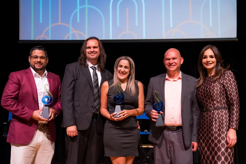 Five honorees stand on stage holding blue and green glass awards.