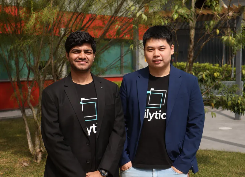 Two men wearing blazers and matching black logo T-shirts stand outdoors.