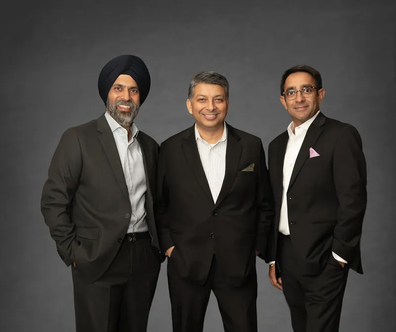 Three people in dark suits stand together against a gray backdrop.