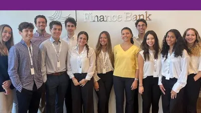 A group of young adults wearing lanyards stands in front of a Finance Bank sign.