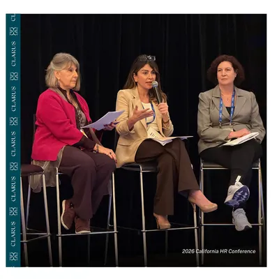 Three people seated on a conference panel, with the center speaker holding a microphone.