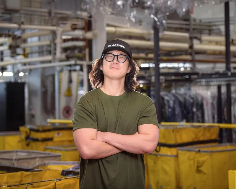 A person in glasses and a black cap stands with arms crossed in an industrial facility.