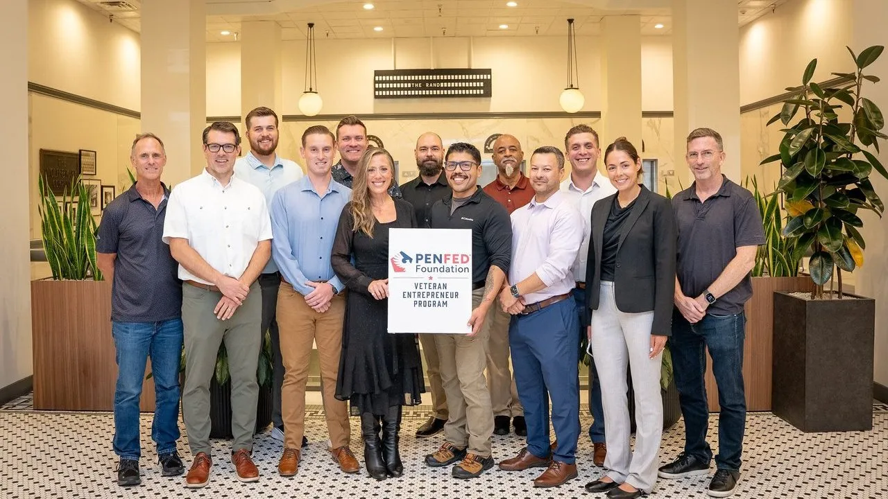 Group of 12 people holding a PenFed Foundation Veteran Entrepreneur Program sign in an office lobby.