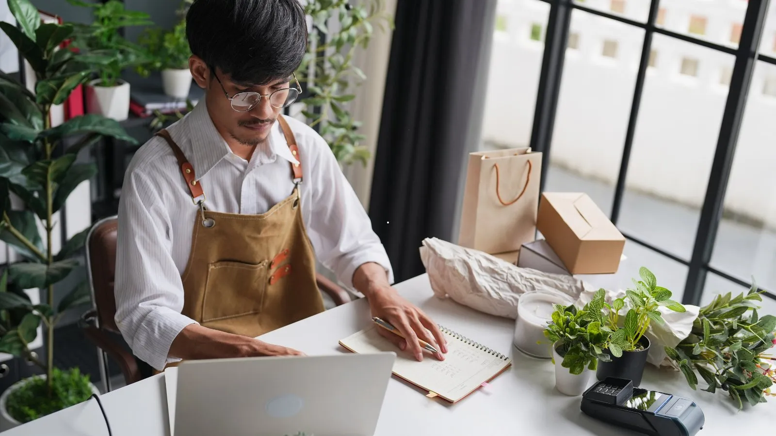 A small shop owner working at a laptop beside parcels and plants